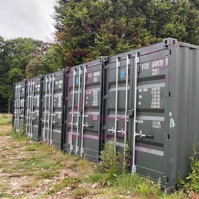 A row of green shipping containers stands in a grassy area, with trees in the background.