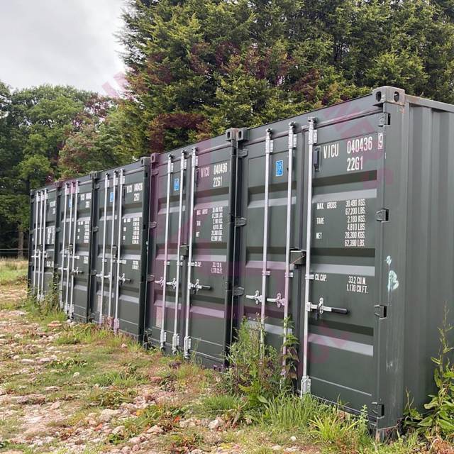 A row of green shipping containers stands in a grassy area, with trees in the background.