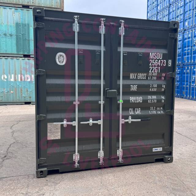 A black shipping container with labels and specs, positioned in a yard with other containers in the background.